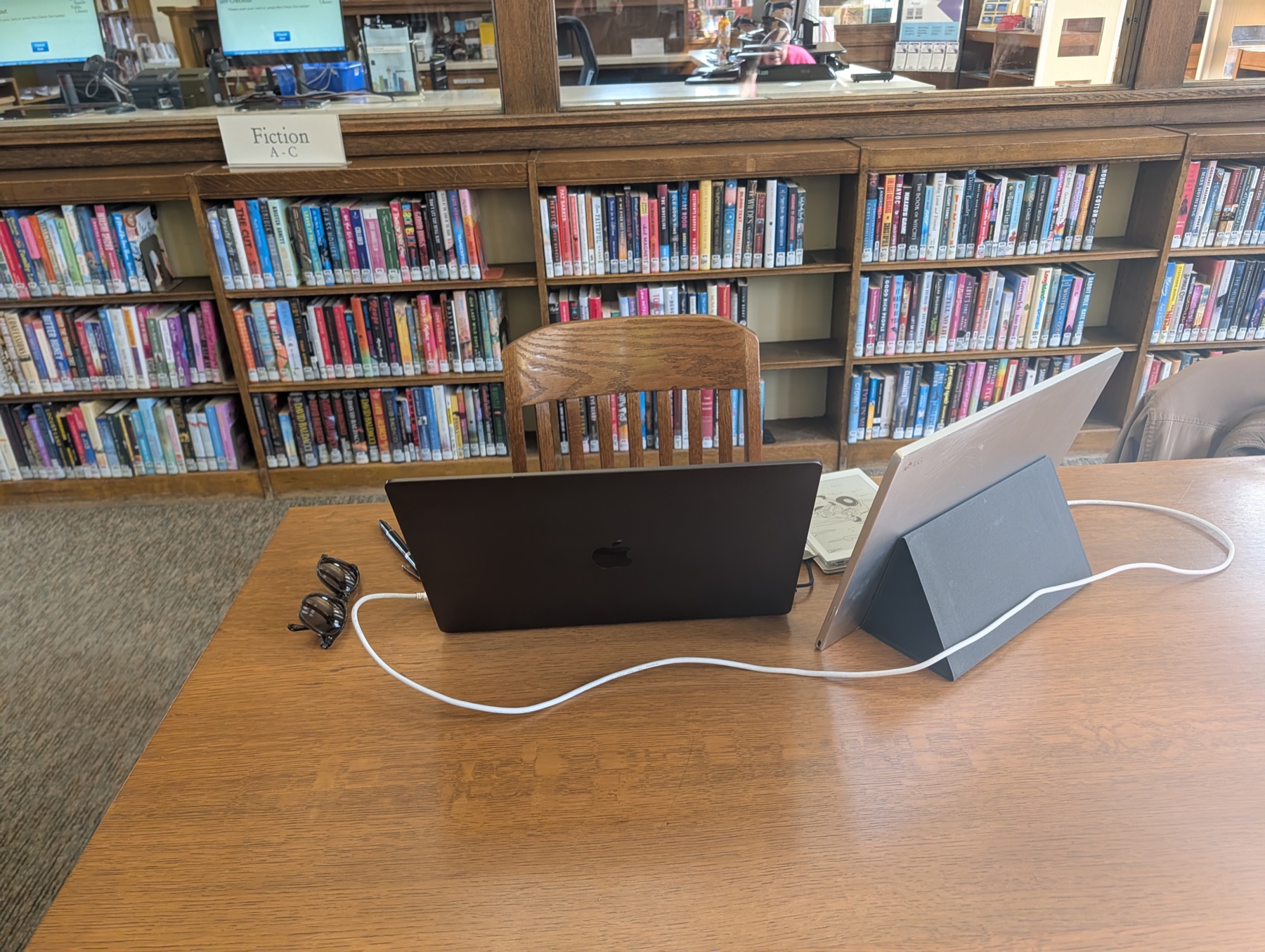A laptop and tablet on a wooden library table in front of bookshelves.